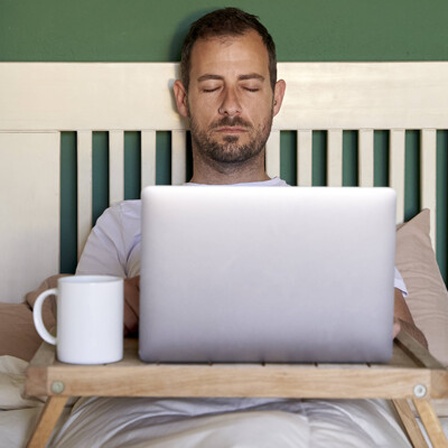 Ein Mann liegt mit geschlossenen Augen im Bett mit Laptop vor sich.