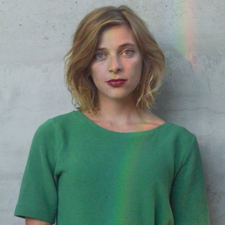 Eine junge Frau steht vor einer grauen Wand und trägt ein grünes T-Shirt.