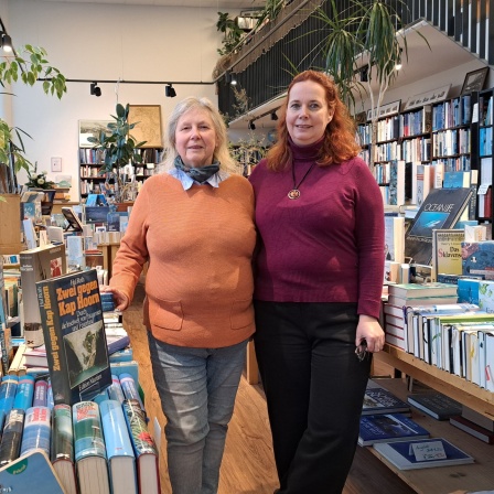 Zwei Frauen stehen in einer Buchhandlung zwischen sehr vielen Büchern.