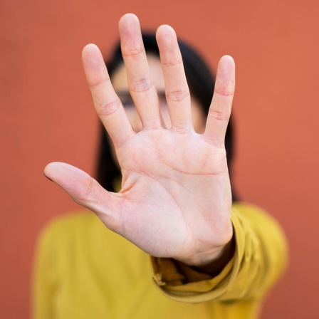 Eine Frau, die in die Kamera blickt und Stopp gebärdet. Das Gesicht ist durch die Hand verdeckt.