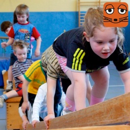 Eine Gruppe sechs Kindern klettert in einer Turnhalle über Kästen und Leitern.