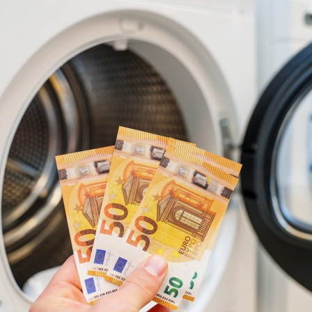 Eine Hand hält drei 50-Euro-Scheine vor eine Waschmaschine.