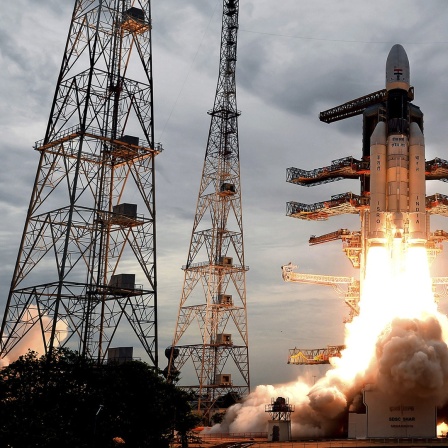 Eine indische Trägerrakete startet ihren Flug ins All Richtung Mond. Um die Rakete herum stehen Masten. Durch den Antrieb der Rakete entstehen Rauchwolken. 