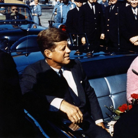 John F. Kennedy sitzt lachend in einer Limousine, neben ihm befindet sich seine Frau Jackie Kennedy.