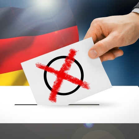 Wahlkarte mit Wahlkreuz wird in eine Wahlurne gesteckt, dahinter eine Deutschlandflagge (Bildmontage)