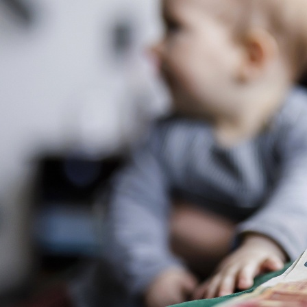 Ein 10-Euro-Schein liegt auf einem Tisch. Im Hintergrund sitzt ein Baby auf dem Schoß einer Frau.