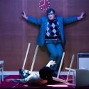 Szene aus dem Theaterstück "humanistää!" am Volkstheater in Wien. Eine Person mit ausgebreiteten Armen, mit Maske steht in einem grauen Anzug mit karriertem Pullunder vor einer brauenen Wand.