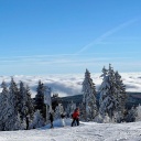 Auf einem Berg liegt viel Schnee. Man sieht auf Höhe des Berges die Wolkendecke und darüber strahlend blauen Himmel und Sonne, die den Schnee hell bescheint. Ein paar Skilangläufer stehen am Berg und blicken auf die Wolken. 