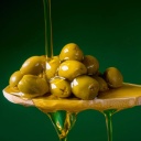 Nahaufnahme von Oliven auf einem Holzlöffel vor grünem Hintergrund. Ein Strahl Olivenöl wird darüber gegossen