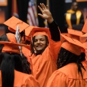Studenten in orangener Kleidung bei der Abschlussfeier. Ein junger Mann reckt einen Arm empor.