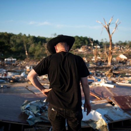 Ein Mann mit schwarzem Cowboyhut steht mit gesenktem Kopf vor den Überresten eines durch Tornados zerstörten Gebäudes.