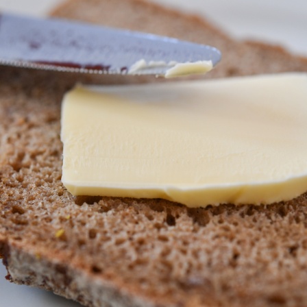 Ein Stück Butter liegt auf einer Scheibe Brot - ein Messer ist daneben zu sehen, um die Butter zu verstreichen.