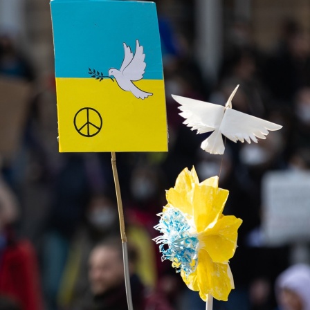 Schilder mit Friedenstaube und Blumen in den Farben der Ukraine werden hochgehalten.