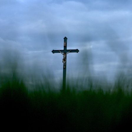 Ein Wegkreuz steht am frühen Morgen unter bewölktem Himmel auf einer Wiese.