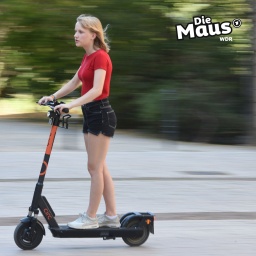 Auf dem Bild ist eine junge Person in einem roten T-Shirt und schwarzen Shorts zu sehen, die auf einem E-Scooter auf einem Bürgersteig fährt. Das "Die Maus"-Logo ist in der oberen rechten Ecke platziert.