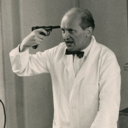 Mann in weißem Kittel mit Fliege hält sich einen Revolver an die rechte Schläfe