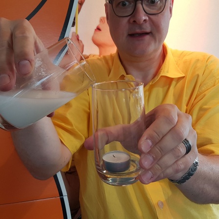 Maus-Wissenschaftsreporter Joachim kippt eine Wasser-Backpulver-Mischung in ein Glas, in dem ein Teelicht steht