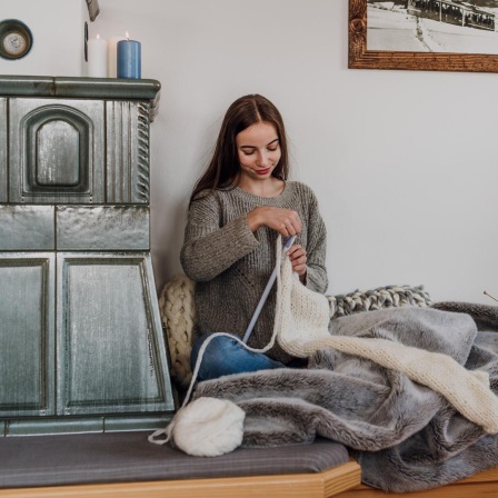 Eine junge Frau sitzt am Kachelofen und strickt.