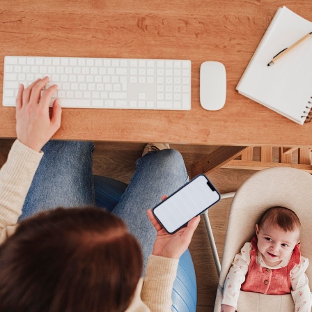 Eine Mutter schaut zugleich ihr Smartphone und ihr Baby in der Liege daneben an, ihre linke Hand liegt auf einer Computertastatur.