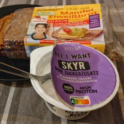 Ein Frühstücksteller mit Mandel-Eiweißbrot der Marke Mestemacher, teilweise noch in der Verpackung, liegt auf einem schwarzen Teller. Daneben steht ein geöffneter Becher Skyr mit einem Löffel darin. Der Skyr ist von der Marke "All I Want", ohne Zuckerzusatz, mit der Geschmacksrichtung Blaubeere-Traube & Lavendel. Die Verpackung weist auf den hohen Proteingehalt und die natürliche Zusammensetzung hin. Alles steht auf einer grauen Tischdecke mit kariertem Muster.
