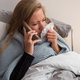 Eine Frau sitzt im Bett mit Taschentusch am Mund und telefoniert