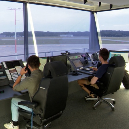 Von hinten sieht man zwei junge Männer in der Kontrollzentrale des Lübecker Flughafen sitzen, die Landebahnen sind ihre Aussicht.
