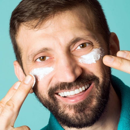 Ein Mann streicht Creme auf seine Augenringe und lacht
