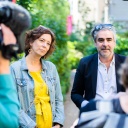 Eva Menasse und Deniz Yücel sprechen im Garten des Literaturhaus Berlin mit Journalisten. Sie trägt ein gelbes Kleid und eine Jeansjacke, er ein helles Hemd und Sakko. Beide haben Brillen ins Haar hochgeschoben. Links ist unscharf ein Mann mit Kamera auf der Schulter von hinten zu sehen, rechts der Hinterkopf und die Schulter einer Frau. 