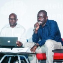 Achille Mbembe und Felwine Sarr sitzen an einem kleinen Tisch, auf dem Schilder mit ihrem Namen stehen. Mbembe hat einen Laptop auf dem Schoß, Sarr spricht in ein Mikrofon.