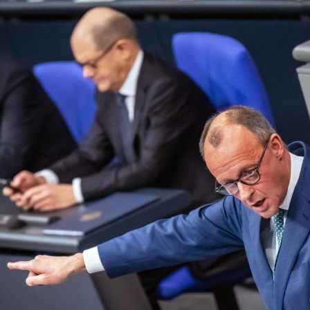 Friedrich Merz spricht im Bundestag. Im Hintergrund schaut Olaf Scholz auf sein Handy.