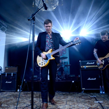 Thorsten Nagelschmidt steht während eines Auftritts mit seiner Band Muff Potter auf der Bühne. Er spielt Gitarre und trägt ein Hemd mit Blumenmuster zum Anzug.