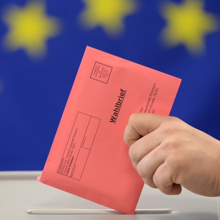 Symbolbild: Vor einer EU-Fahne steht eine Wahlurne, in die eine Person einen Wahlbrief einwirft.