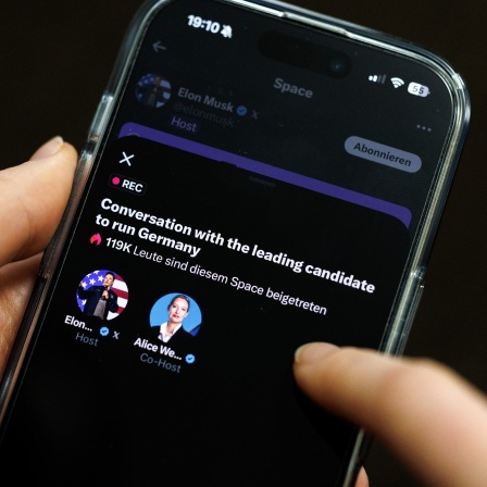 Eine Frau hält ein Smartphone in den Händen, auf dem der Livestream vom Gespräch von Elon Musk mit Alice Weidel auf der Plattform X mit dem Titel "Conversation with the leading candidate to run Germany“ läuft.