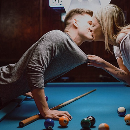Paar küsst sich über einem Billiardtisch.