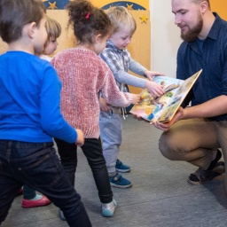 Ein Erzieher im Kindergarten bespricht mit Kindern zur musikalischen Früherziehung ein Buch über Zootiere.