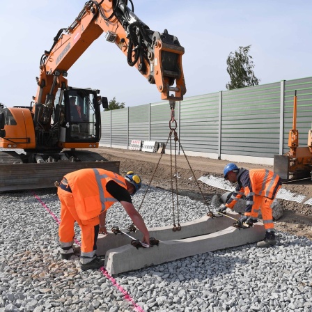 Arbeiter verlegen mit Hilfe eines Raupenbaggers Betonschwellen.