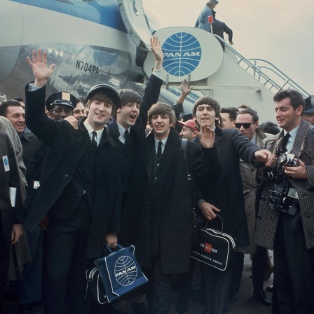 John Lennon winkend, Paul McCartney, Ringo Starr and George Harrison stehen vor einem Jumbo (AP Photo), im Hintergrund die Treppe, die sie vom Flugzeug herabgegangen sind. Die vier Musiker sind umgeben von Männern, rechts steht ein Fotograf.