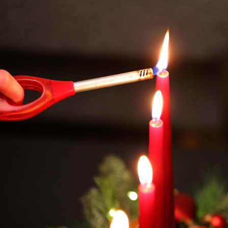 Kerzen auf einem Adventsgesteck werden mit einem langen Feuerzeug angezündet.