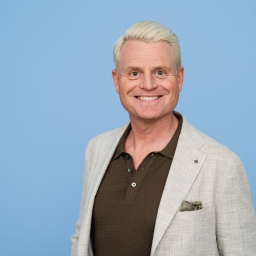 Der Moderator Guido Cantz steht vor einem blauen Hintergrund und lächelt in die Kamera.