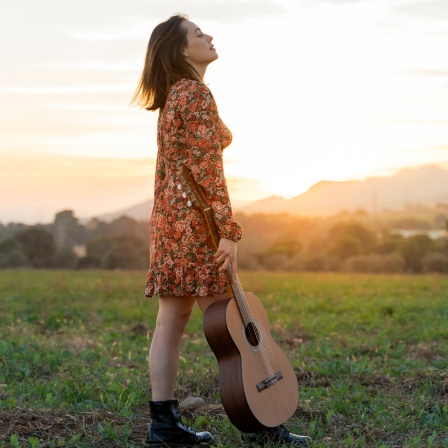 Junge Frau steht mit geschlossenen Augen bei Sonnenuntergang auf einer Wiese, dabei hält sie eine Gitarre.