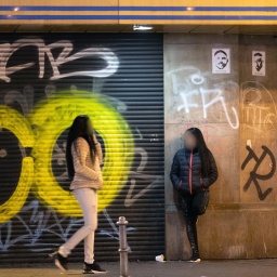 Zwei Prostituierte stehen im Frankfurter Bahnhofsviertel vor einem mit einem geschlossenen Torrolladen,  der ebenso wie die angrenzende Wand mit Graffiti-Tags besprüht ist.