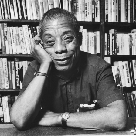 James Baldwin sitzt, seinen Kopf auf die Hand gestützt, lächelnd vor einem Bücherregal