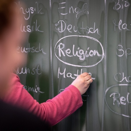 Eine Lehrerin schreibt mit Kreide einen Stundenplan mit Religionsunterricht an eine Tafel.