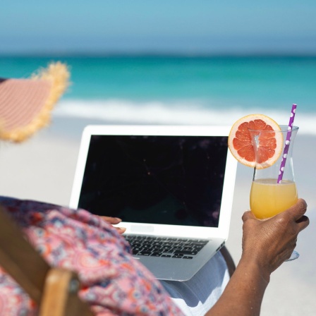 Frau mit Sonnenhut sitzt in einem Liegestuhl am Strand, sie hält ein Getränk in der Hand und hat ihren Laptop auf dem Schoß.