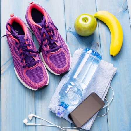 Sportschuhe, Handtuch, Wasserflasche Handy mit Kopfhörern und Obst auf einem blau gestrichenen Holzboden. Symbolbild
