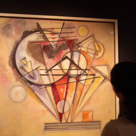 Zu sehen ist ein abstraktes Bild, das Wassily Kandinsky gemalt hat. Wesentliche Elemente darin sind Kreise und Dreiecke. Der Hintergrund des Kunstwerks ist in Beigetönen gehalten. Das Hauptmotiv erscheint in den Farben Rot, Orange, Gelb und Violett.