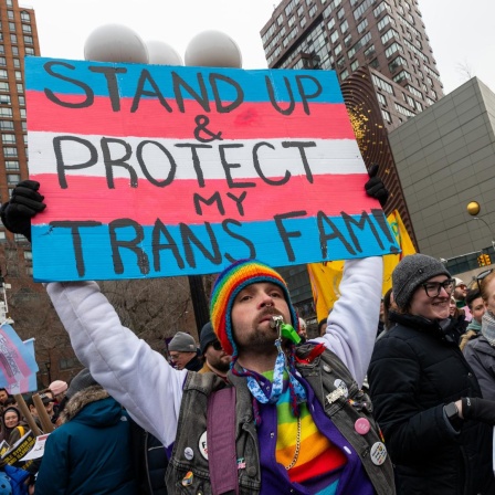 Eine Person mit vielen Regenbogenaccessoires an der Kleidung, einer Sonnenbrille und einem Bart, hält ein Schild hoch, auf dem vor den Farben der Transgender-Pride-Fahne steht „Stand Up & Protect my Trans Fam!“.