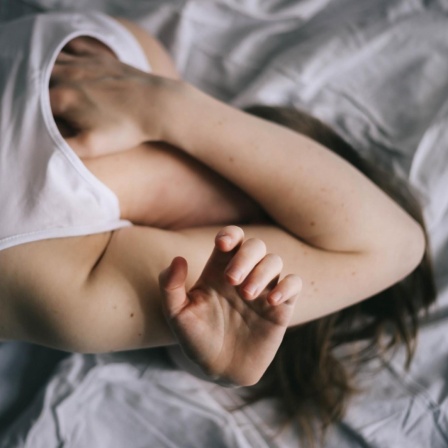 Eine Frau liegt im Bett und verbirgt das Gesicht unter ihren Armen.