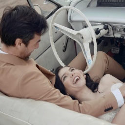 Glückliche lächelnde Frau, die in einem weißen Kleid, vermutlich einem Hochzeitskleid, auf den Knien ihres vermutlich frisch vermählten Mannes im Cabrio liegt. Er sitzt am Steuer und hält liebevoll ihre Hand.
