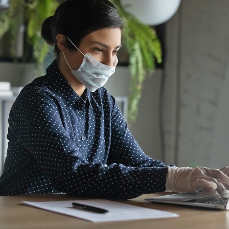 Eine junge Frau arbeitet mit Schutzmaske und Handschuhen an einem Laptop